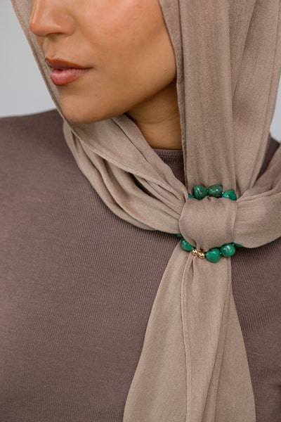 Close-up of a beige hijab with emerald green hoop on a neutral background.